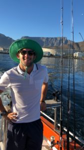 The image depicts a person on a boat engaged in recreational fishing, with rods lined up in the background. They are dressed in a white polo, beige shorts, and a green hat, set against a scenic backdrop of water and mountains under a clear sky. The individual’s face is concealed for privacy. This setting suggests leisure or tourism activities in a picturesque outdoor location.