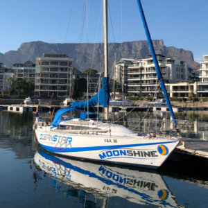 A sailboat named ‘MOONSHINE’ is docked in tranquil waters, with a backdrop of buildings and a mountain under a clear blue sky, suggesting favorable sailing conditions. The scene captures the serene atmosphere of a marina, ready for seafaring adventures